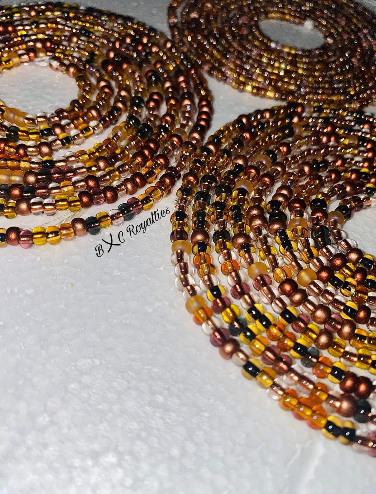 Multiple beaded necklaces in brown and black on a textured surface with 'BXC Royalties' branding.