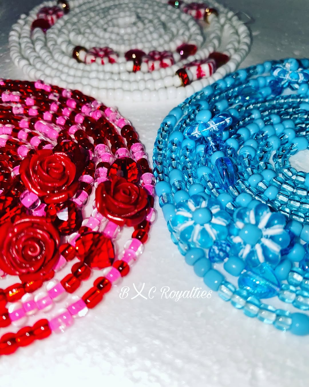 Three beaded waist-beads  in red, pink, and blue on a white background with 'BXC Royalties' branding.