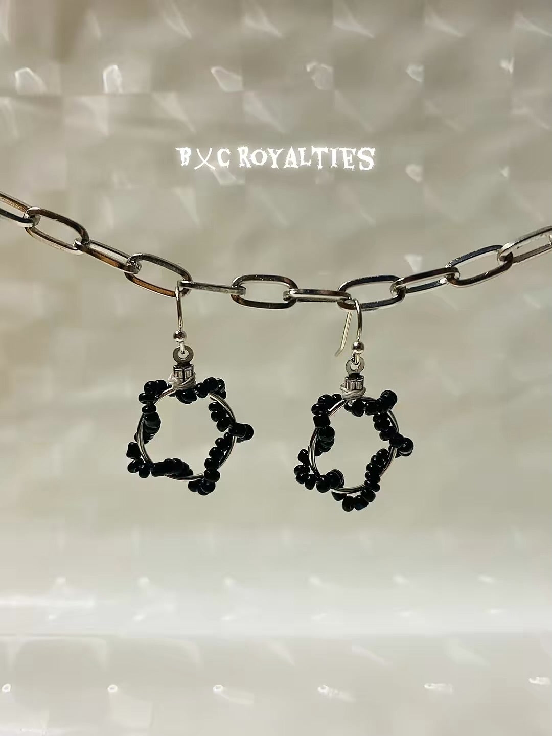 Black star-shaped earrings hanging from a chain against a neutral background with 'BxC Royalties' branding.