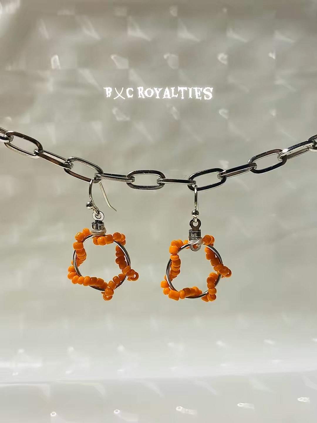 Pair of orange beaded earrings on a chain with 'BXC Royalties' branding.