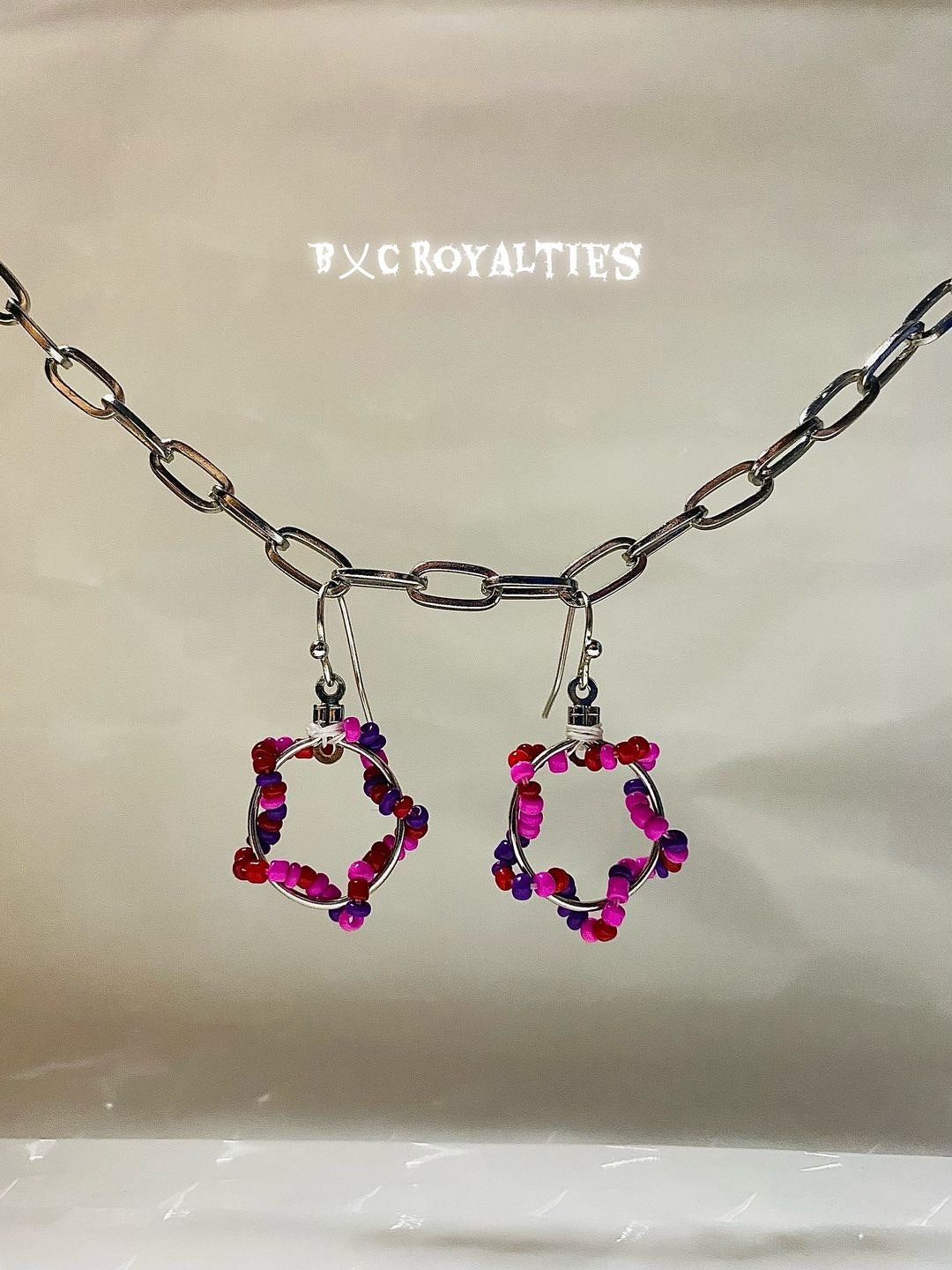 Pair of hoop earrings with pink and purple beads on a beige background, branded 'BXC Royalties'.