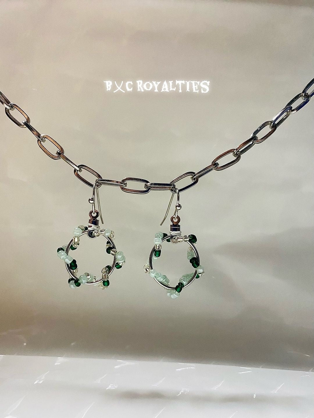 Earrings with green gemstones on a beige background, featuring the brand 'B&C Royalties'.