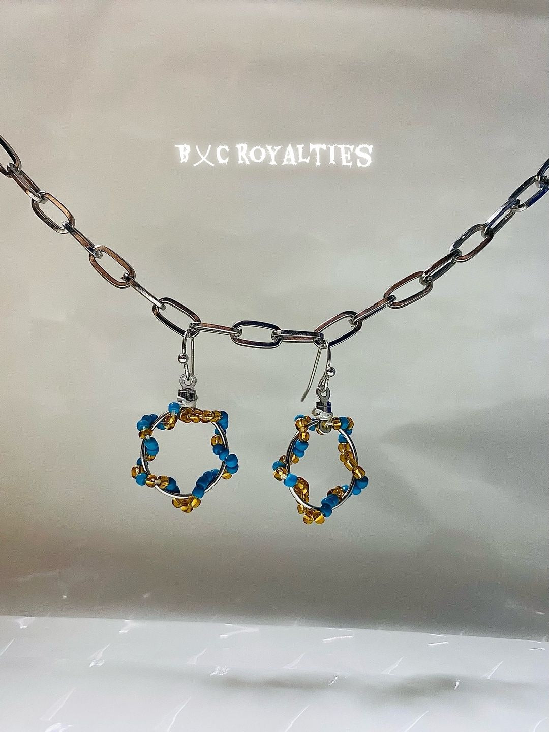 Pair of earrings with blue and gold design hanging from a chain on a reflective surface.