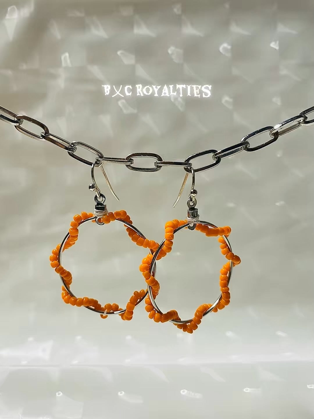 Hoop earrings with orange beads on a chain against a textured background with 'BXC Royalties' branding.