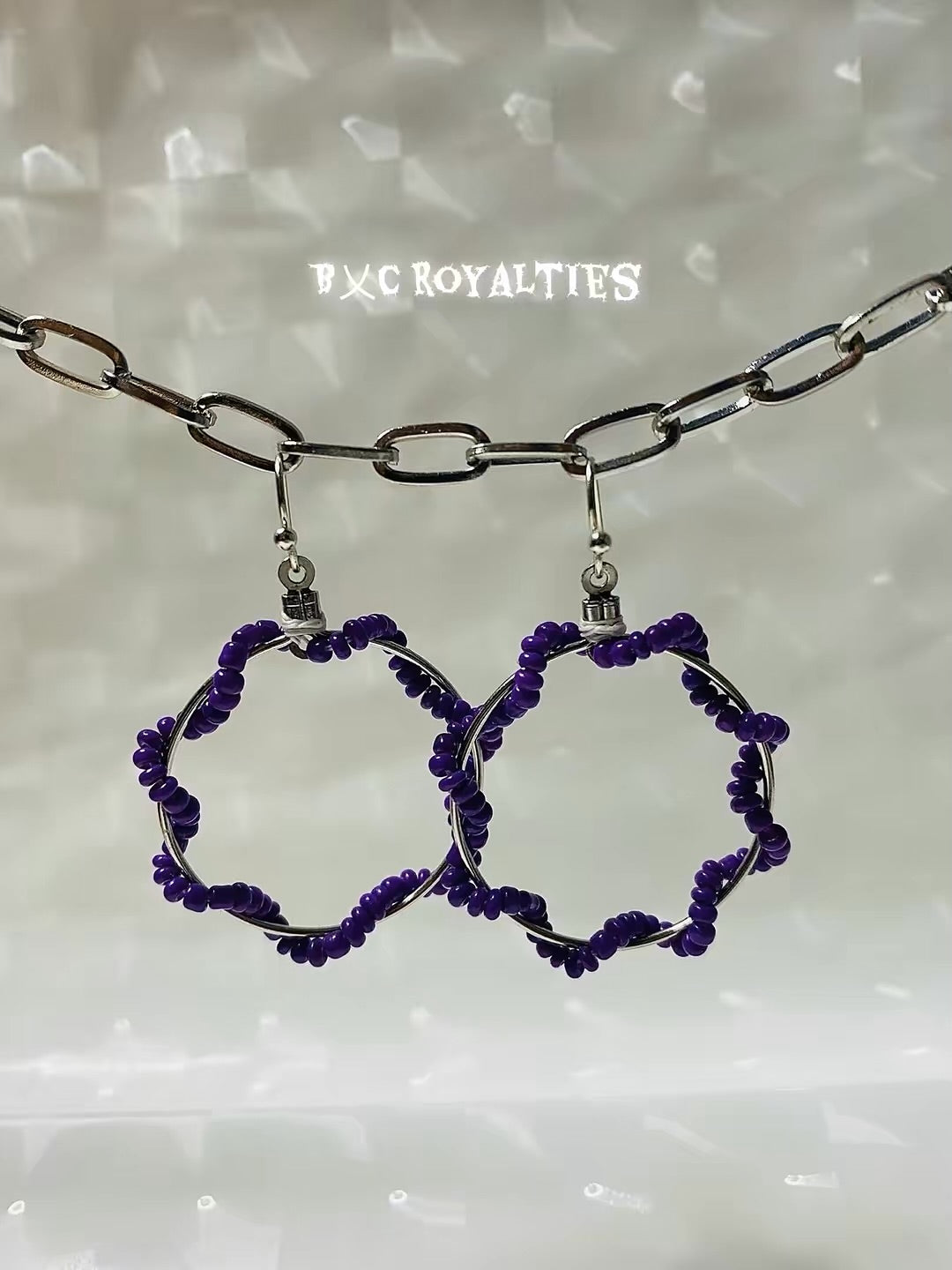 Purple beaded hoop earrings on a chain against a textured white background with 'BXC Royalties' branding.