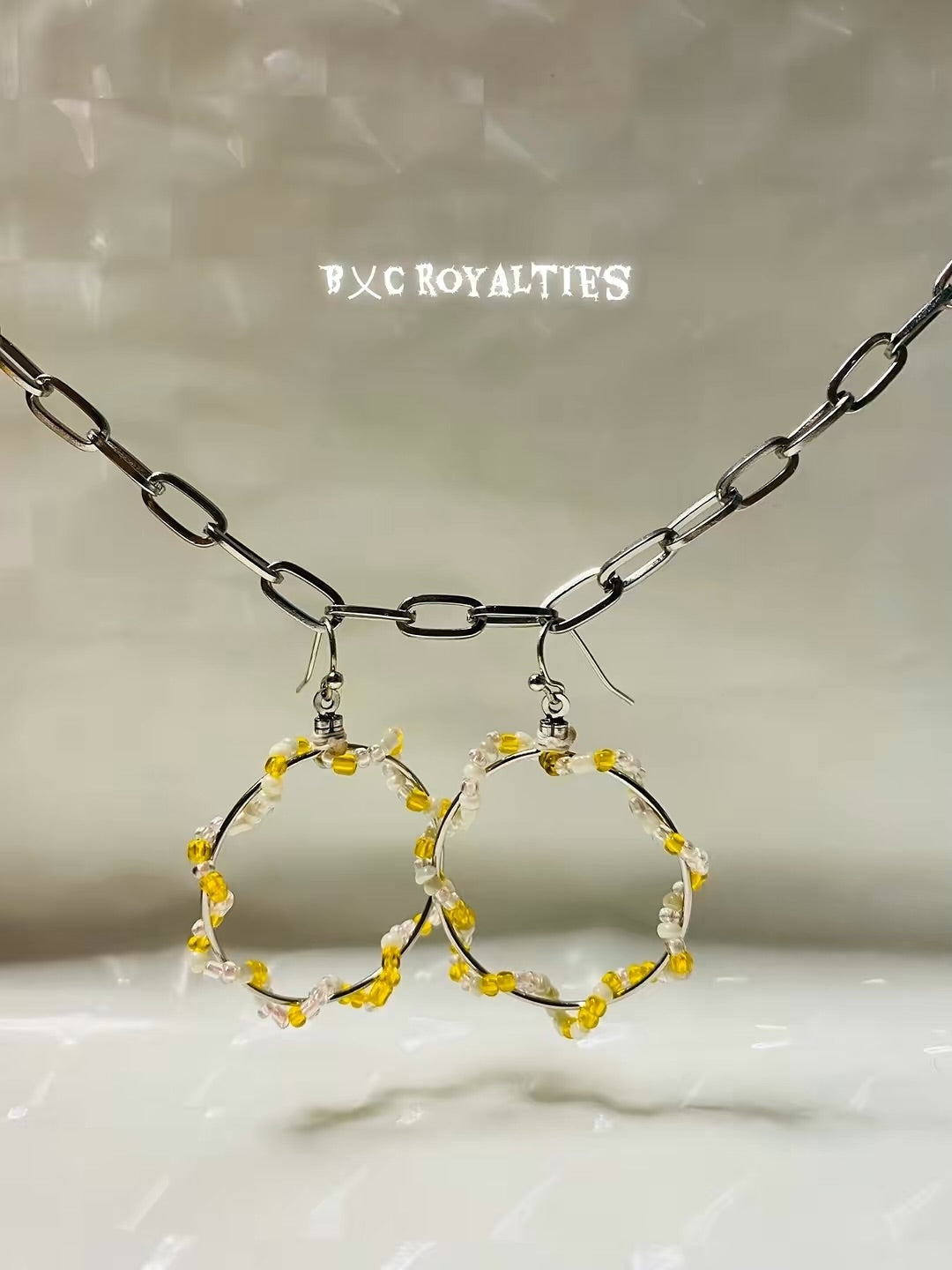 Hoop earrings with yellow and white accents on a neutral background, featuring the brand 'BXC Royalties'.