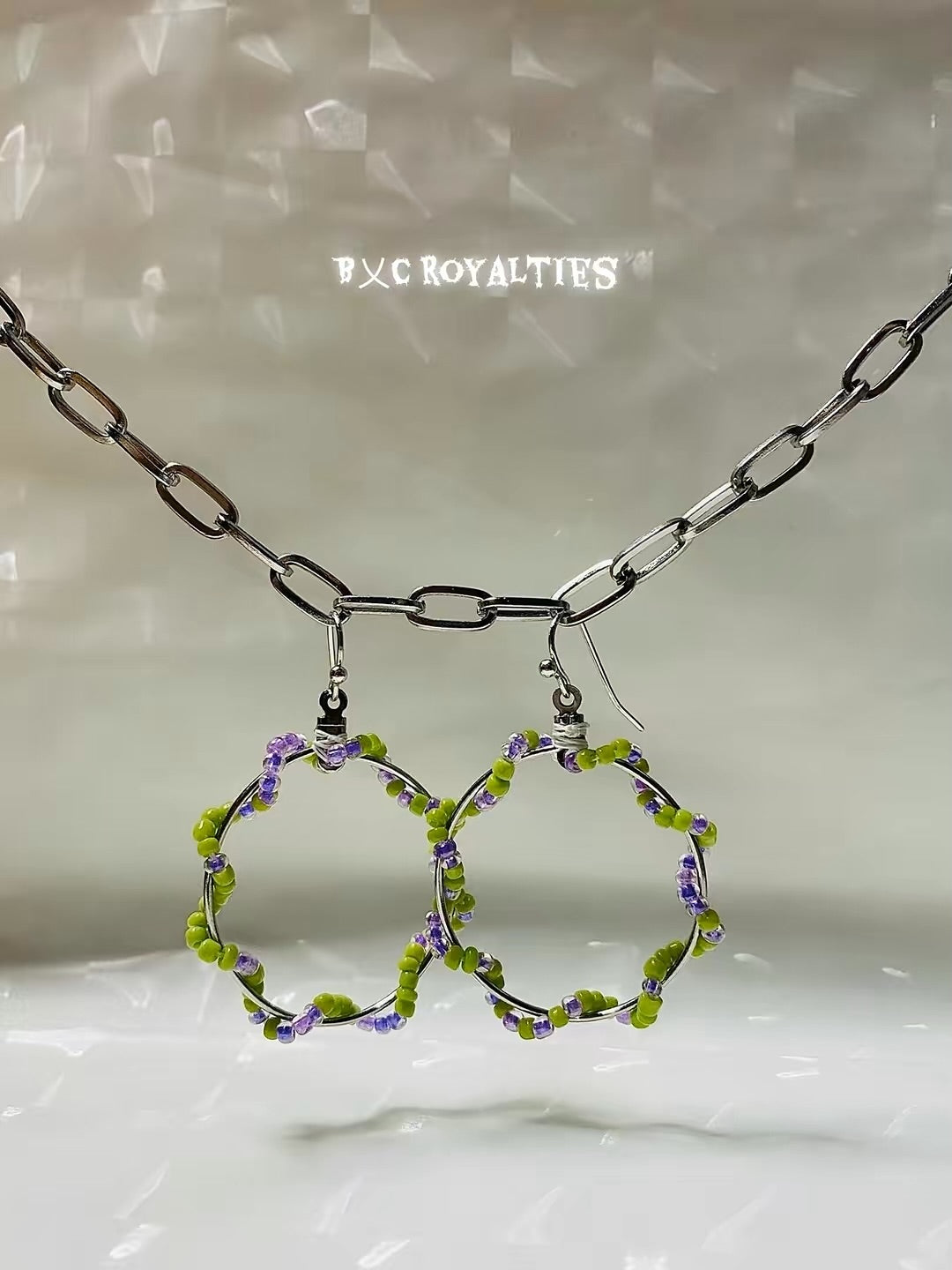 Pair of hoop earrings with green and purple beads on a reflective surface, branded 'BXC Royalties'.