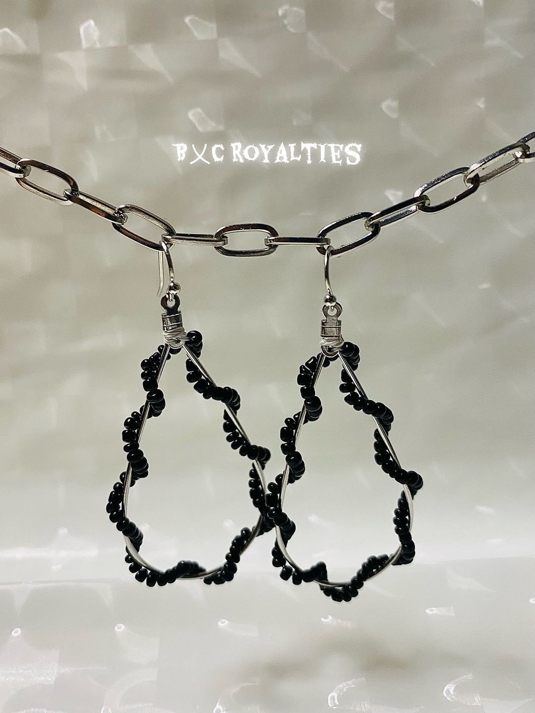 Black teardrop-shaped earrings on a chain against a neutral background with 'BxC Royalties' branding.