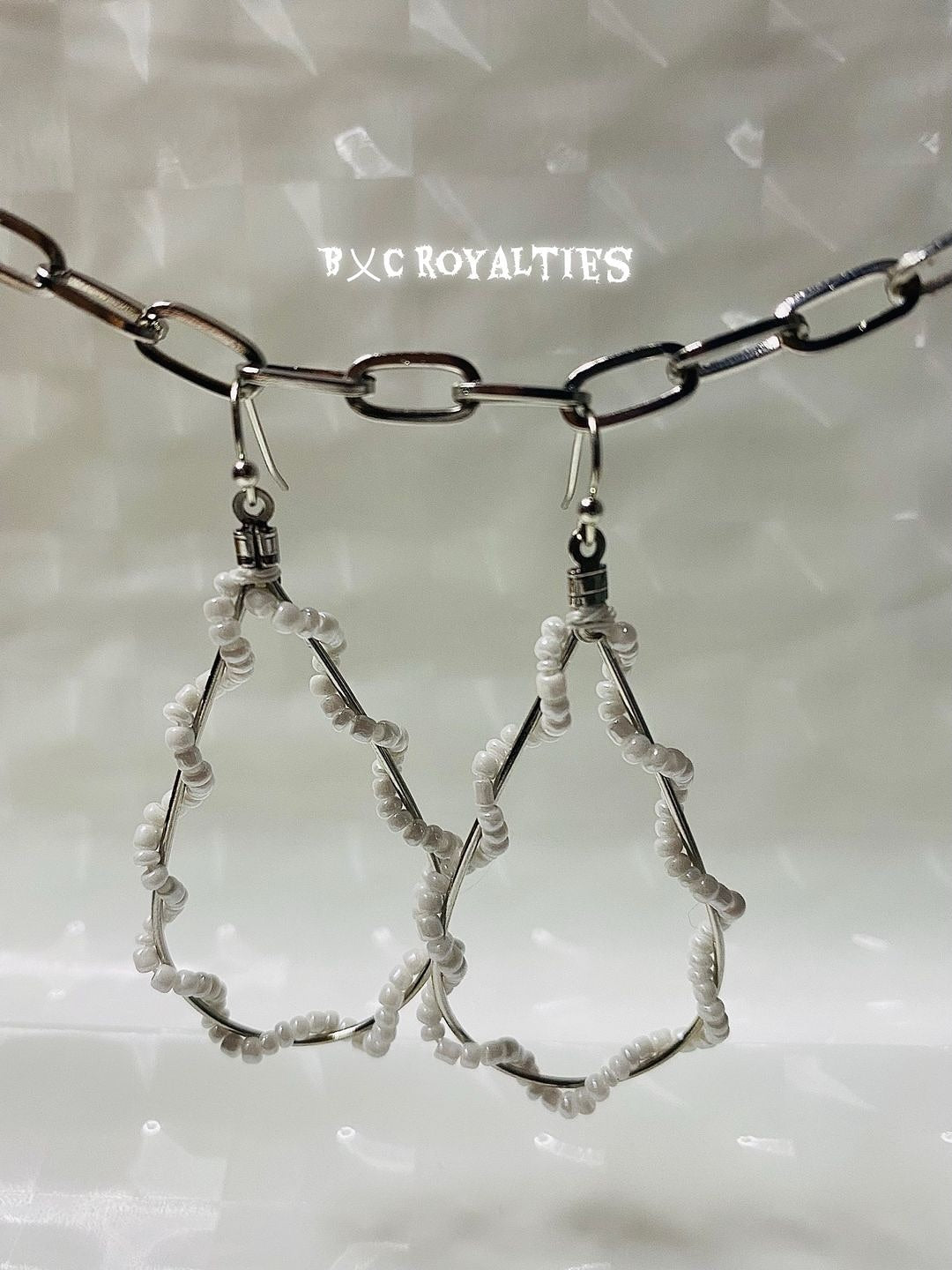 Teardrop-shaped earrings with a chain on a textured white background, featuring the brand 'BXC Royalties'.