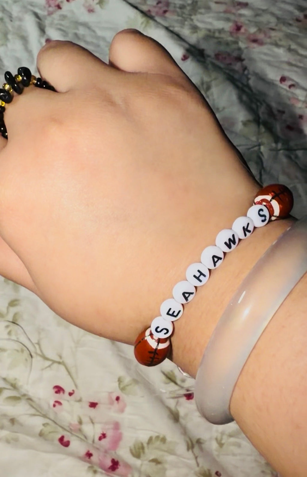 Hand wearing a bracelet with 'SEAHAWKS' beads on a floral fabric background