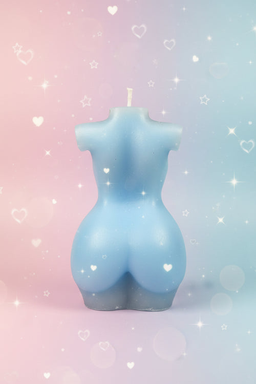 A light blue body-shaped candle with a molded torso and short sleeves, standing upright on a round table against a textured beige wall.