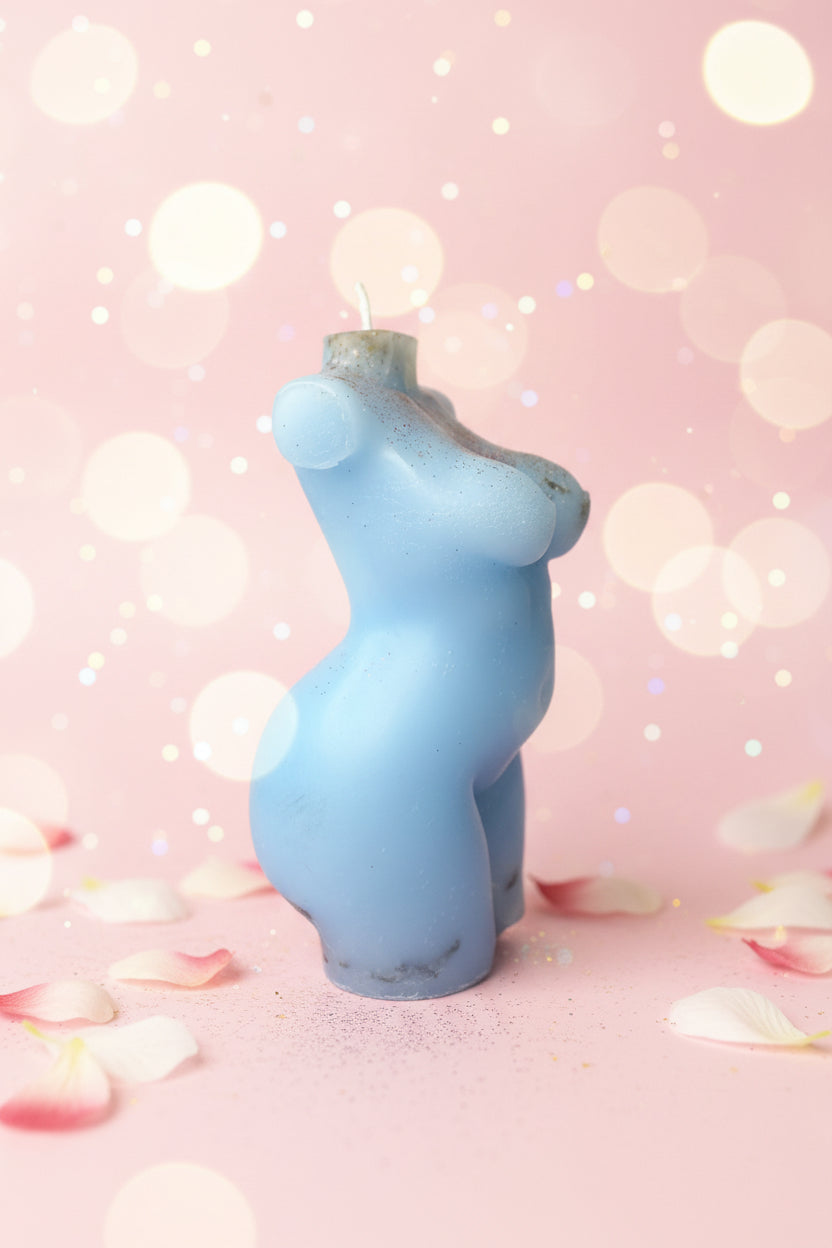 A light blue sculptural candle shaped like a stylized torso, shown standing upright on a round surface with a textured beige wall behind it.