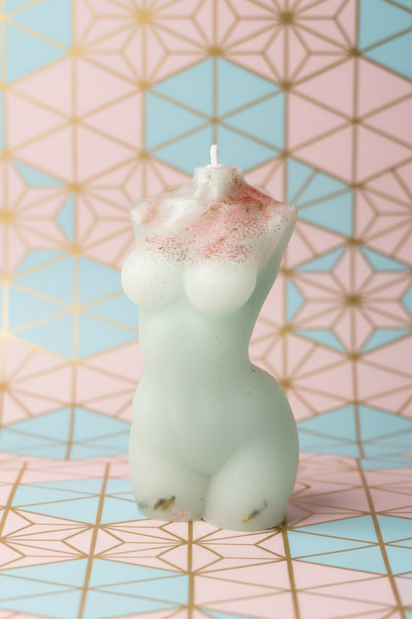 A small sculpted candle shaped like a stylized figure, shown in a pale translucent off-white color with pink speckled detailing near one end. The candle is photographed against a textured off-white wall with a blue and red surface at the right edge.
