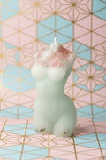 A small sculpted candle shaped like a stylized figure, shown in a pale translucent off-white color with pink speckled detailing near one end. The candle is photographed against a textured off-white wall with a blue and red surface at the right edge.