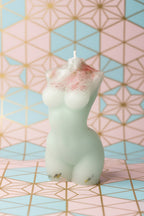 A small sculpted candle shaped like a stylized figure, shown in a pale translucent off-white color with pink speckled detailing near one end. The candle is photographed against a textured off-white wall with a blue and red surface at the right edge.