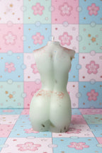 A white silicone or rubber novelty candle shaped like a female torso, shown against a textured beige wall with a blue object on the right.