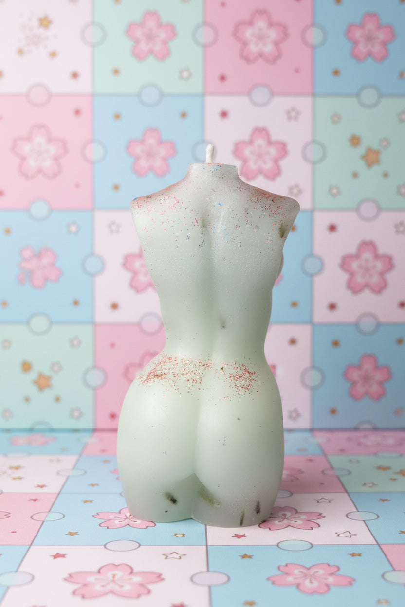 A white silicone or rubber novelty candle shaped like a female torso, shown against a textured beige wall with a blue object on the right.
