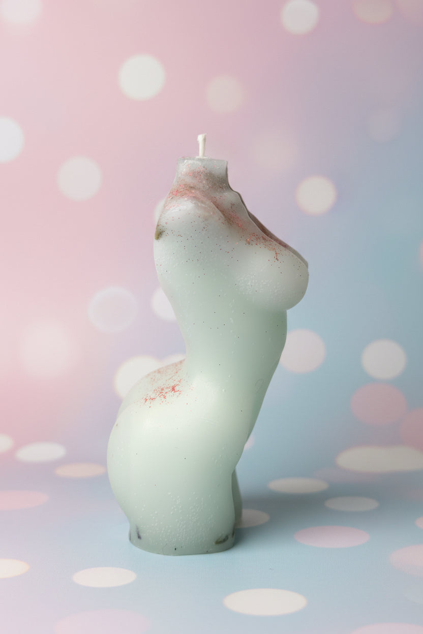 A novelty sculptural candle shaped like a stylized female torso, made of frosted translucent white glass with small red speckles, shown against a textured beige wall and a blue/red background element.
